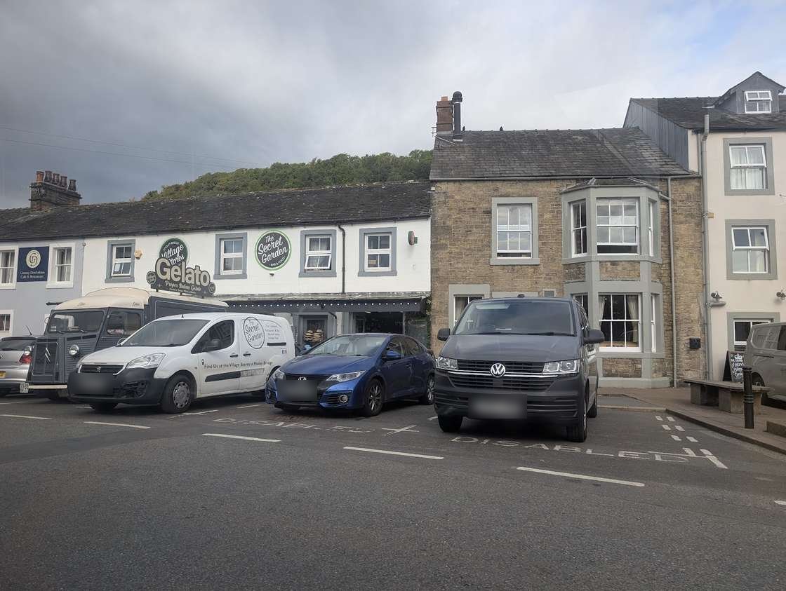 Cars and vans parked in front of whitewashed and stone buildings in Pooley Bridge, a gelato cafe sign among them, a wooded hill behind under a grey sky