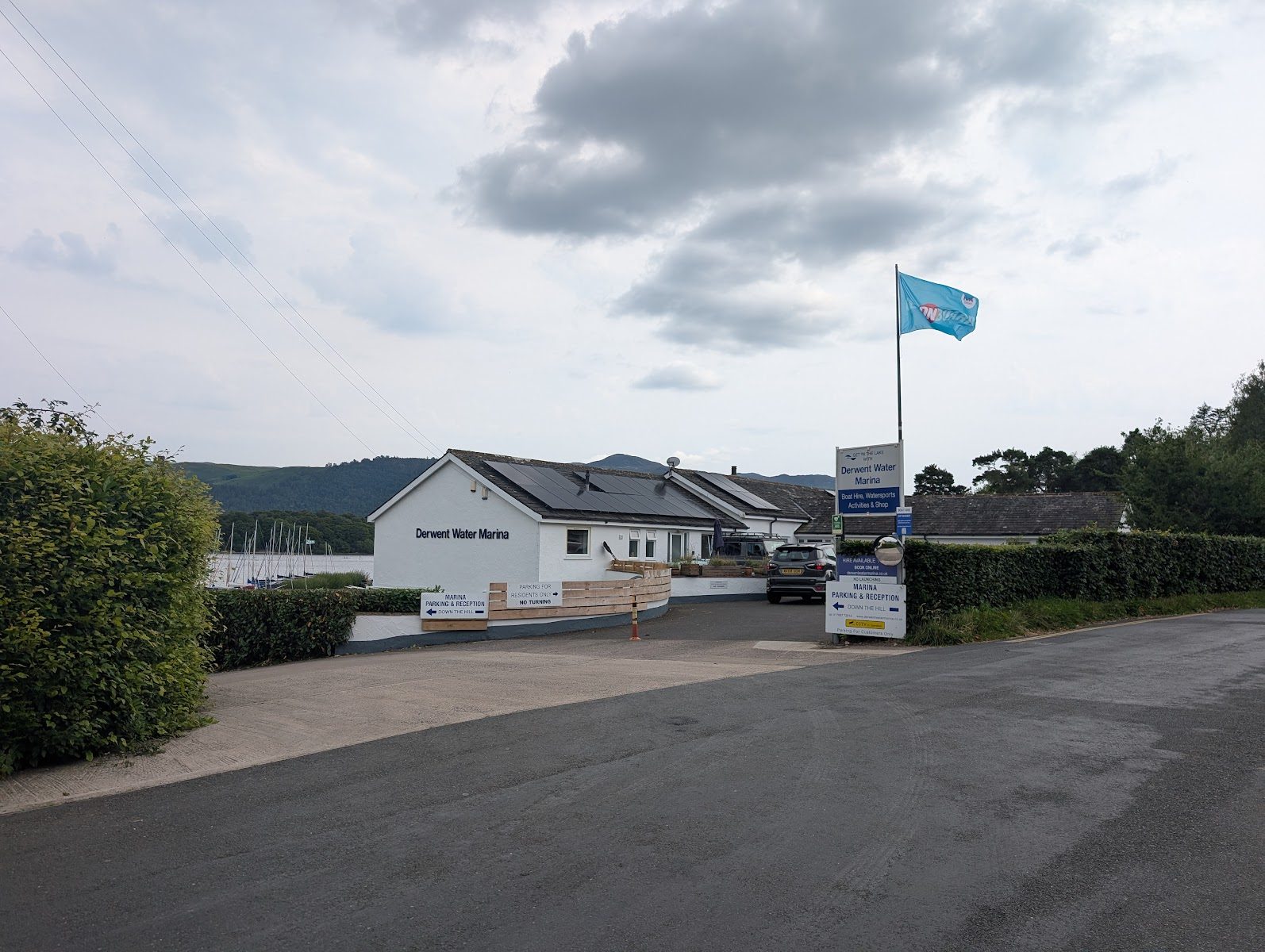 The entrance to Derwent Water Marina at Portinscale: a white single-storey building with solar panels, parking and reception signs, a flag, and boat masts beyond the hedge