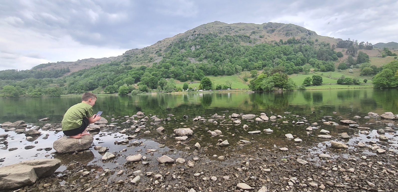 A boy crouching on a rock at the stony edge of Rydal Water, the calm lake reflecting a wooded, bracken-covered fell under grey cloud