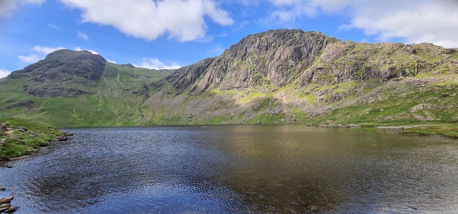Stickle Tarn below the great rock face of Pavey Ark, Harrison Stickle's rounded top on the left, green slopes and a bright, cloud-dotted sky