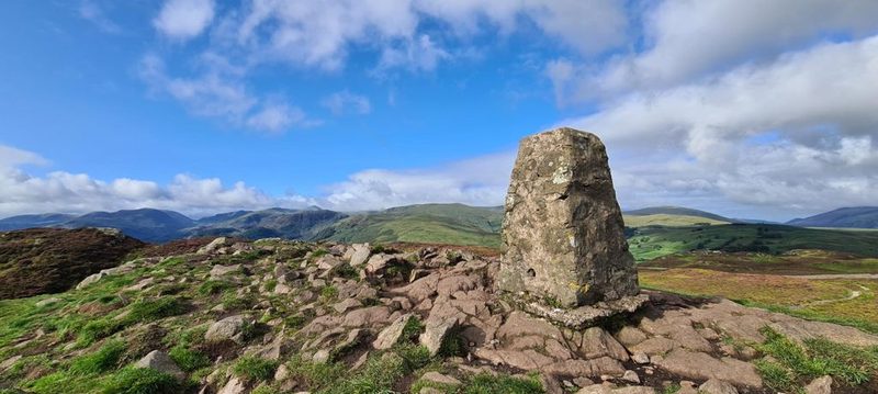 The weathered stone trig pillar on the rocky summit of Gowbarrow Fell, with heather and outcrops around it and a long line of fells beyond