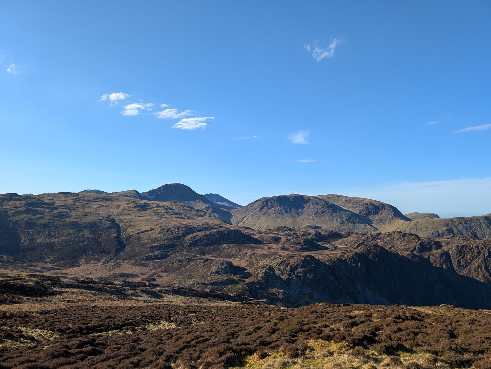 A wide sweep of dark heather and rocky knolls with a high line of crag-topped fells beyond, under a cloudless blue sky