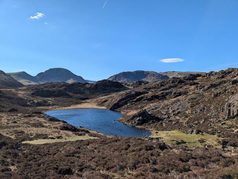 A small dark tarn among heather and rock outcrops, with a shapely pointed fell and a long craggy ridge on the skyline beyond