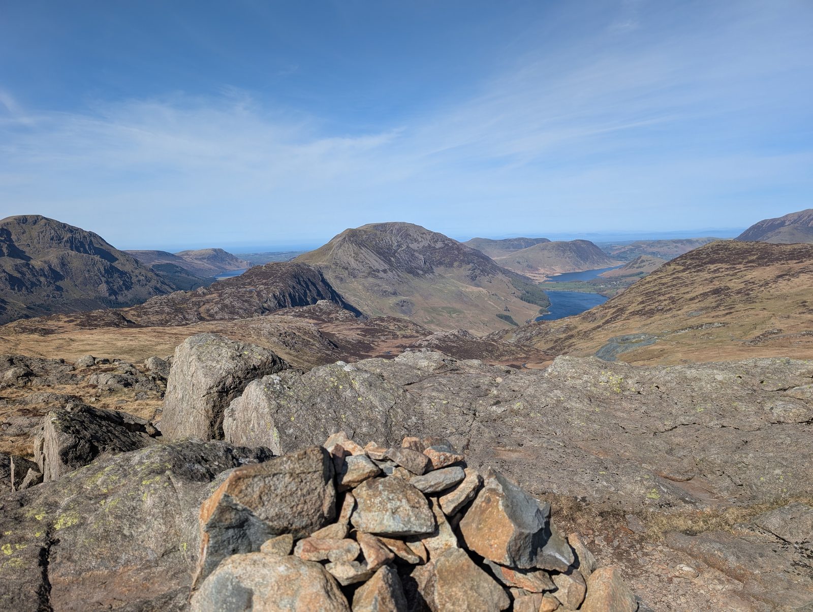 A cairn on bare rock slabs looking out over a rocky top to Crummock Water and Buttermere lying in their valleys far below