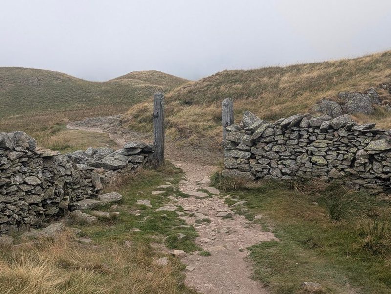 A gap between two drystone walls with old wooden gateposts still standing, a stony path running through it onto rolling grass tops with cloud on the skyline