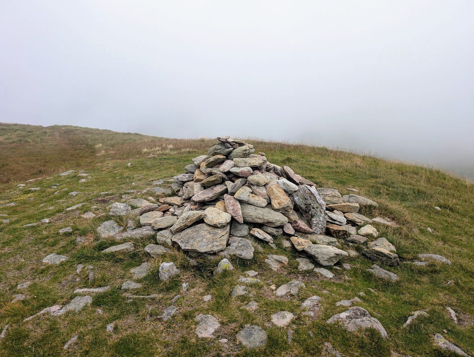 A large, loosely built cairn of flat slabs on a grassy summit, with thick white cloud closing in just beyond it
