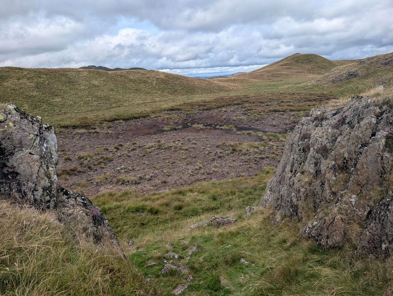 A rocky notch on a heathery ridge looking across a wide peaty hollow to smooth grass tops beyond, under a sky of broken cloud