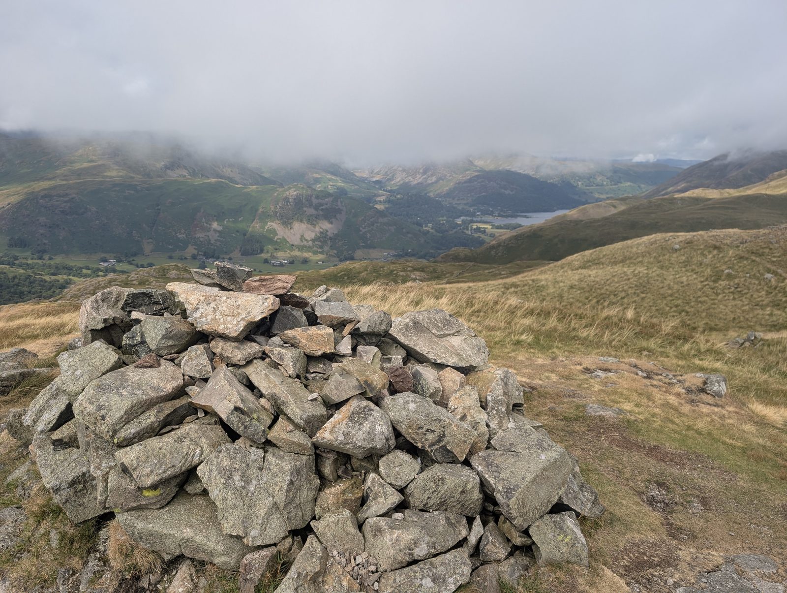A big cairn of angular blocks on a grassy top, looking down over the wooded valley and a glimpse of Ullswater with cloud on the fells beyond