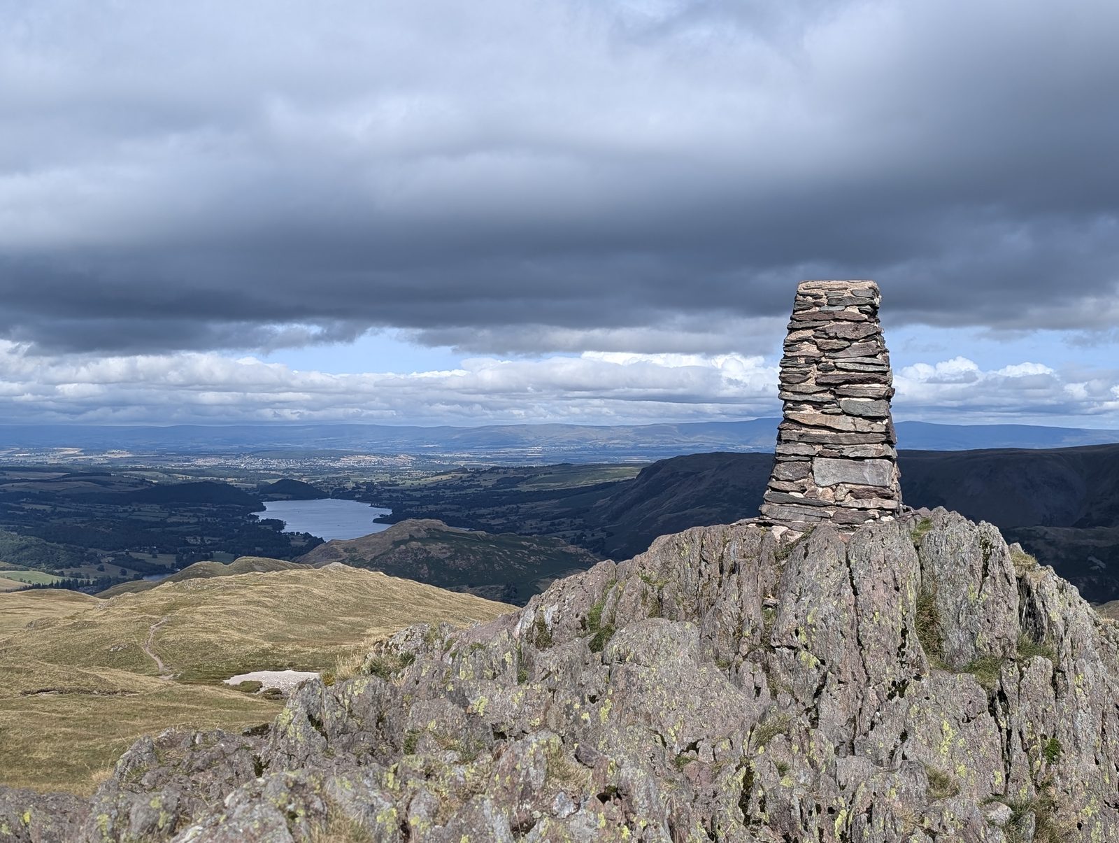 A tall drystone column on a rocky summit, looking out over Ullswater and the green lowlands under a heavy bank of cloud