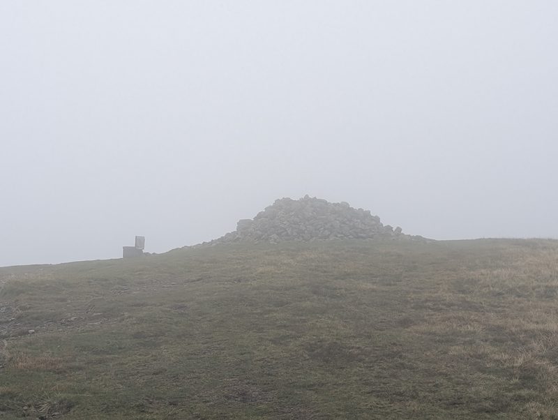 The big pile of stones on the top of High Pike in mist, with the summit bench to its left