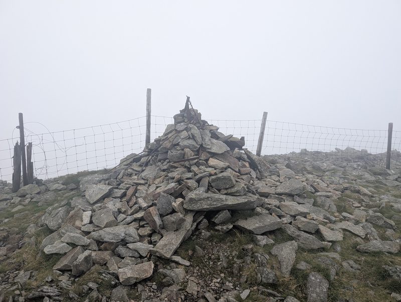 The summit cairn on Great Calva, built against a wire fence on stony ground, in mist