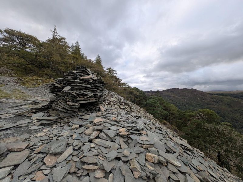 A cairn built entirely of stacked slate slabs on a huge spoil heap of loose slate, with larch trees behind and the valley beyond