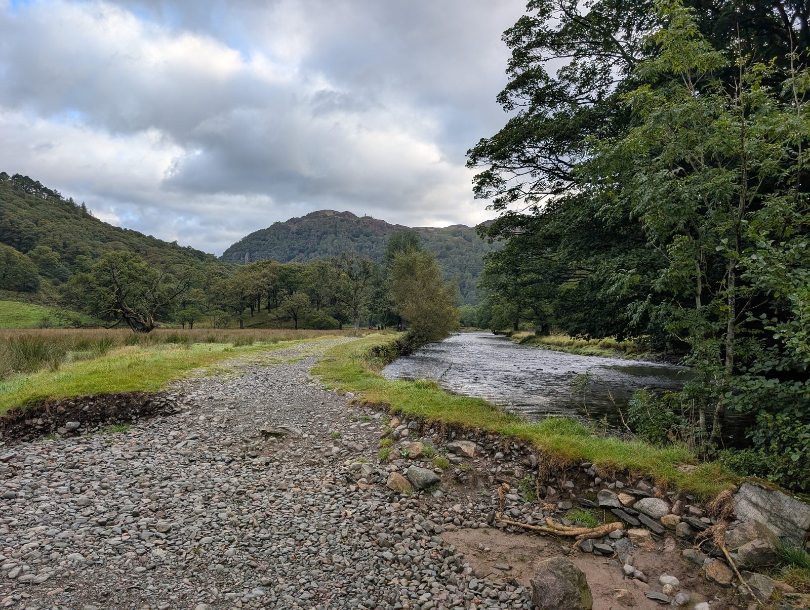 A wide gravel path beside a clear river running through a wooded valley, with a small wooded crag rising ahead beyond the meadows