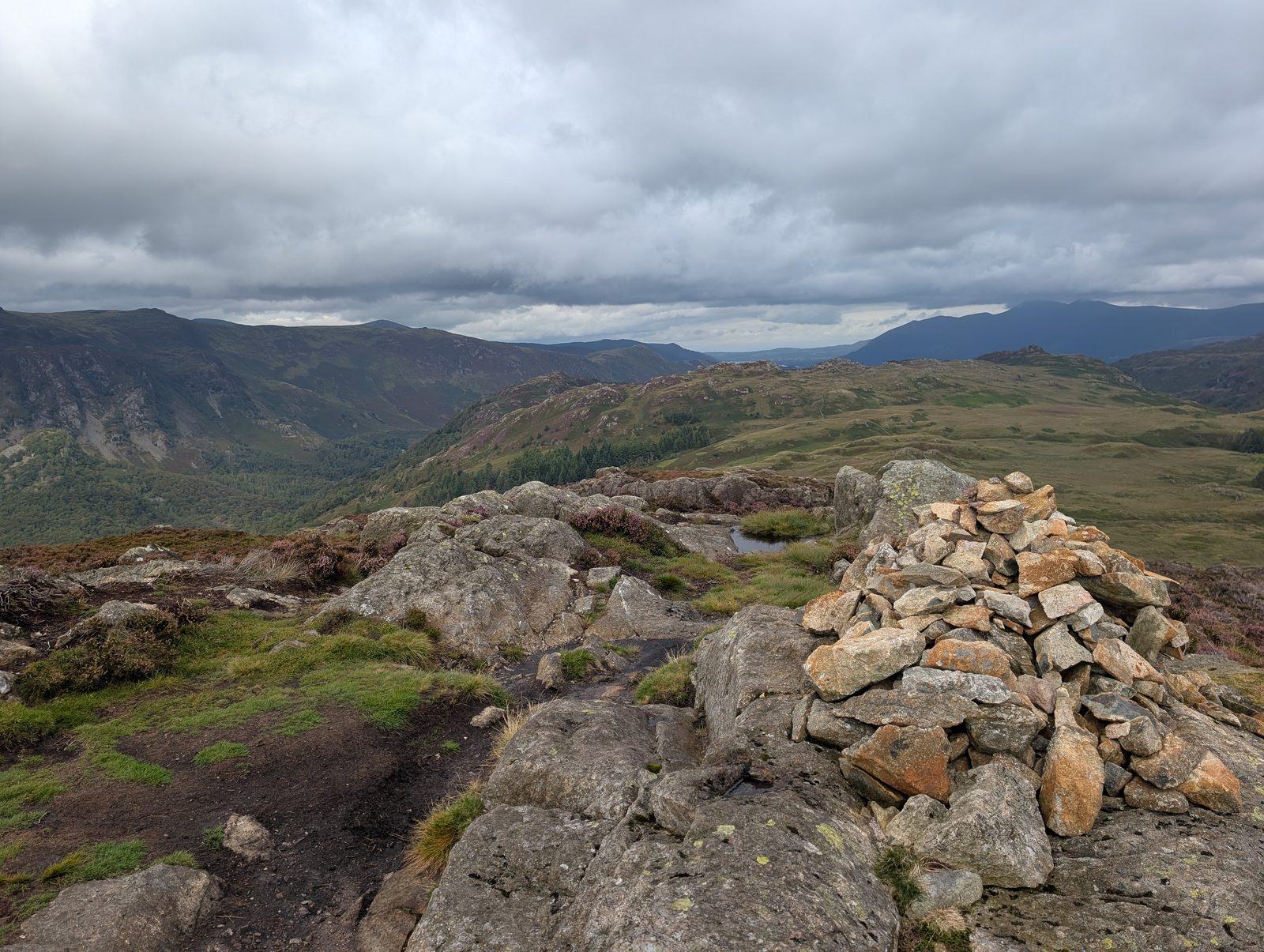 A cairn of pale angular blocks on a rocky summit, with heather, a small pool and a wooded valley falling away to the left