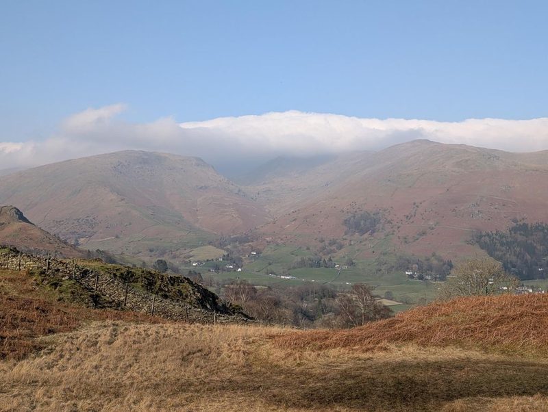 A drystone wall on a bracken-covered shoulder, looking across a green valley of walled fields and farms to a long ridge with cloud lying flat along its top
