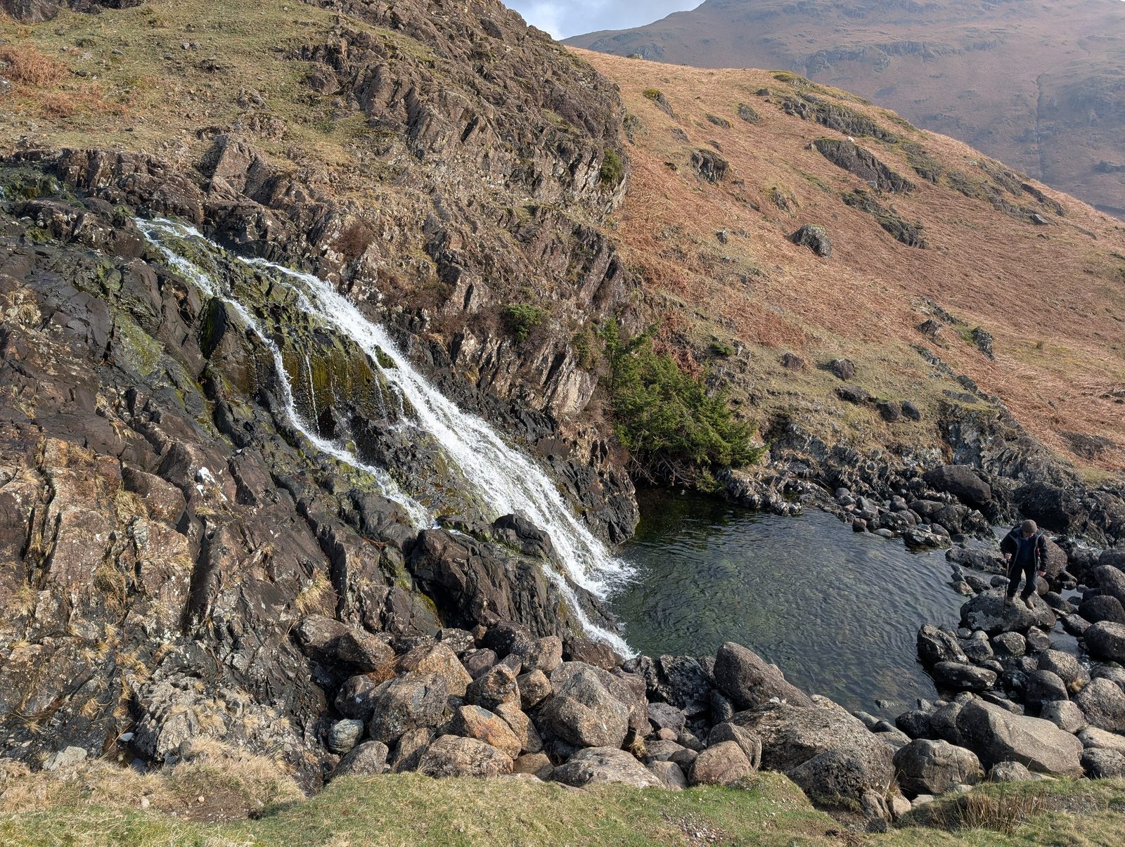 A waterfall spilling down a mossy rock face into a clear pool, with a walker picking a way over the boulders at its edge