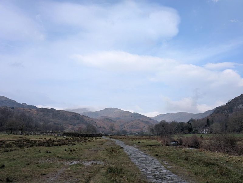 A pitched stone causeway path crossing flat valley pasture towards low wooded fells, with a drystone wall and a white house among trees