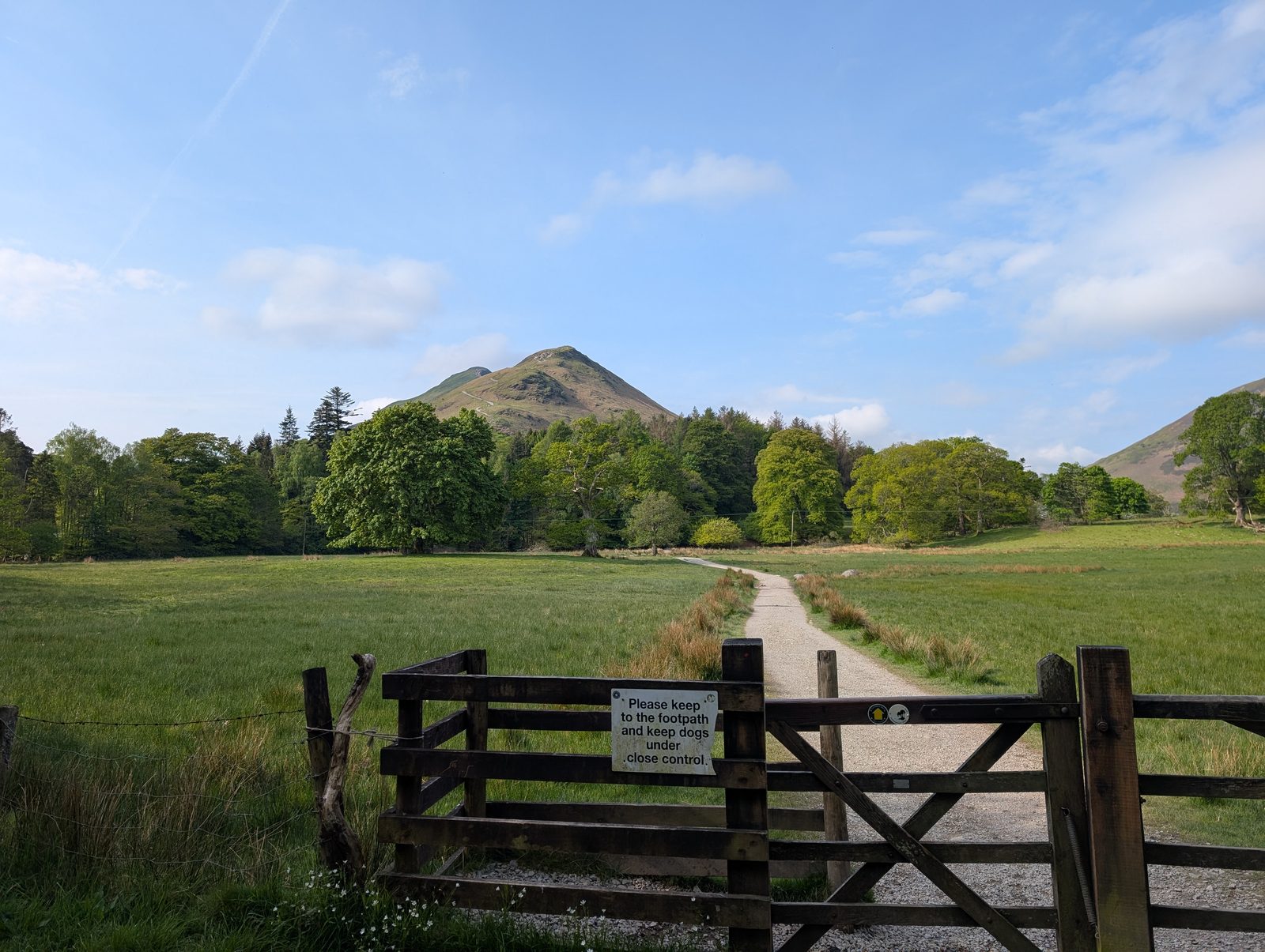 Cat Bells rising as a shapely green cone beyond a meadow and a five-bar gate, with a notice asking walkers to keep to the footpath