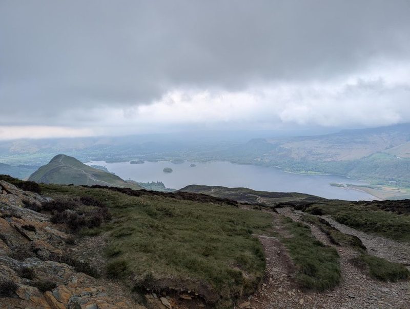 Derwent Water and its islands below a rain-heavy sky, with the ridge falling away and a worn stony path running down in the foreground