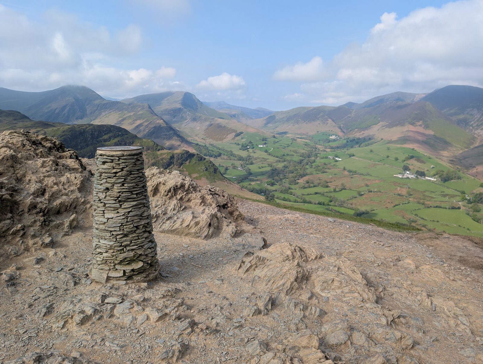 The drystone-built trig column on Cat Bells' bare, slate-strewn summit, with the green Newlands valley and its farms below and fells all around
