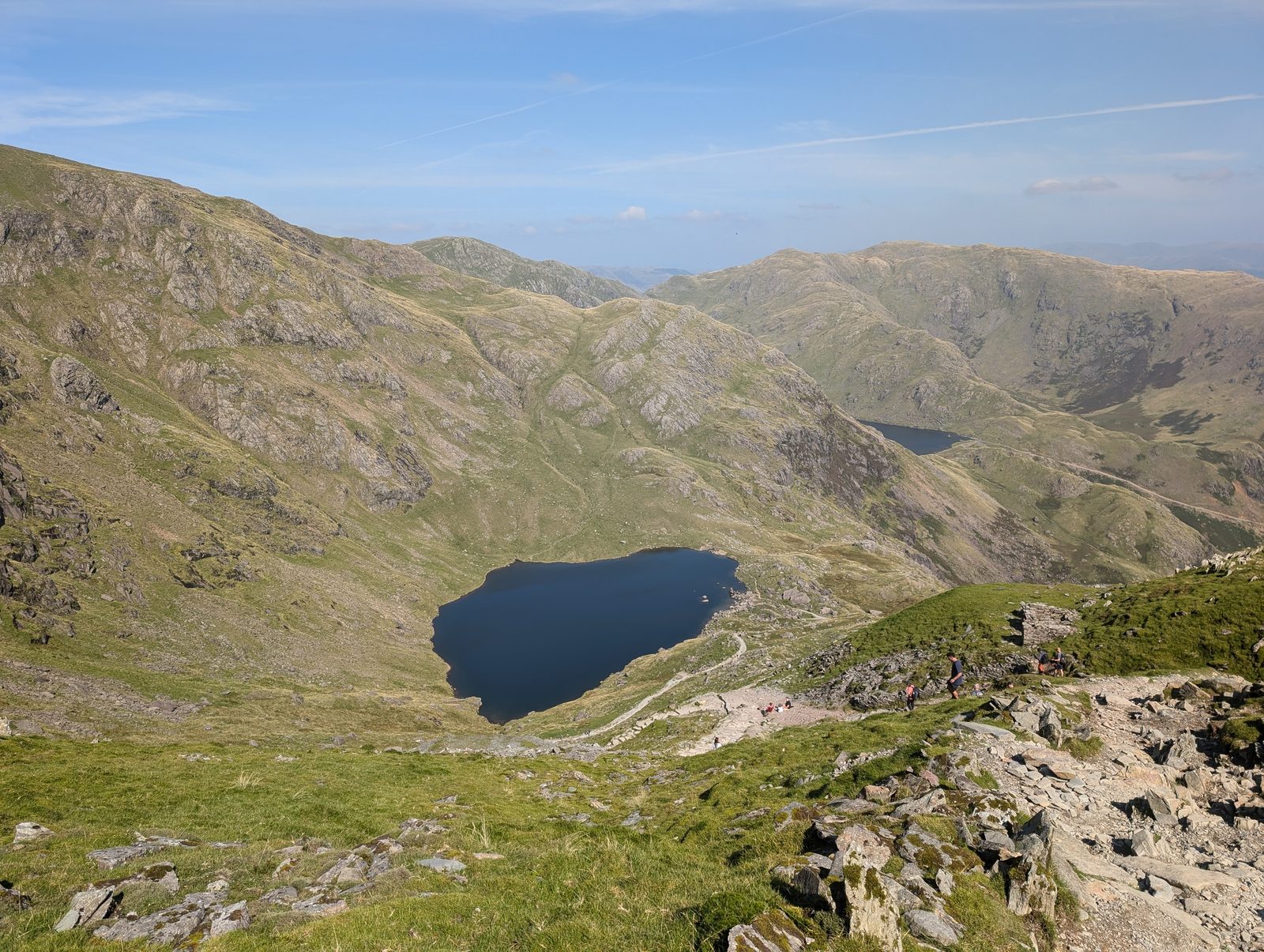 Looking down from high on the climb to the dark tarn of Low Water in its corrie, with walkers strung out along the stony path and a second tarn beyond