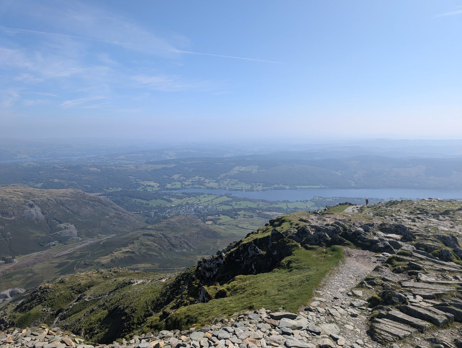 A stony path along the summit ridge with the fellside dropping away to Coniston village and the long stretch of Coniston Water in the haze below
