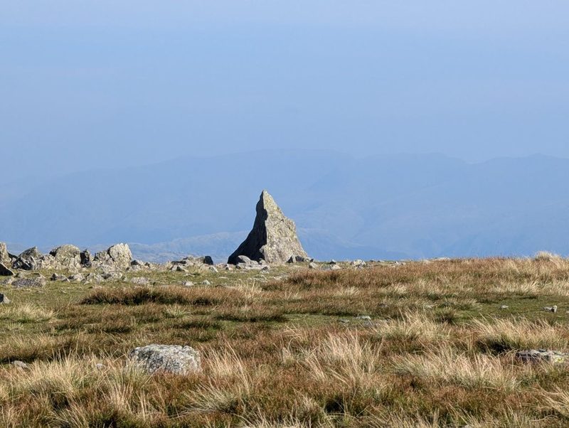 A tall pointed slab of rock standing alone on a plateau of tussocky grass, with range after range of hazy blue fells behind it