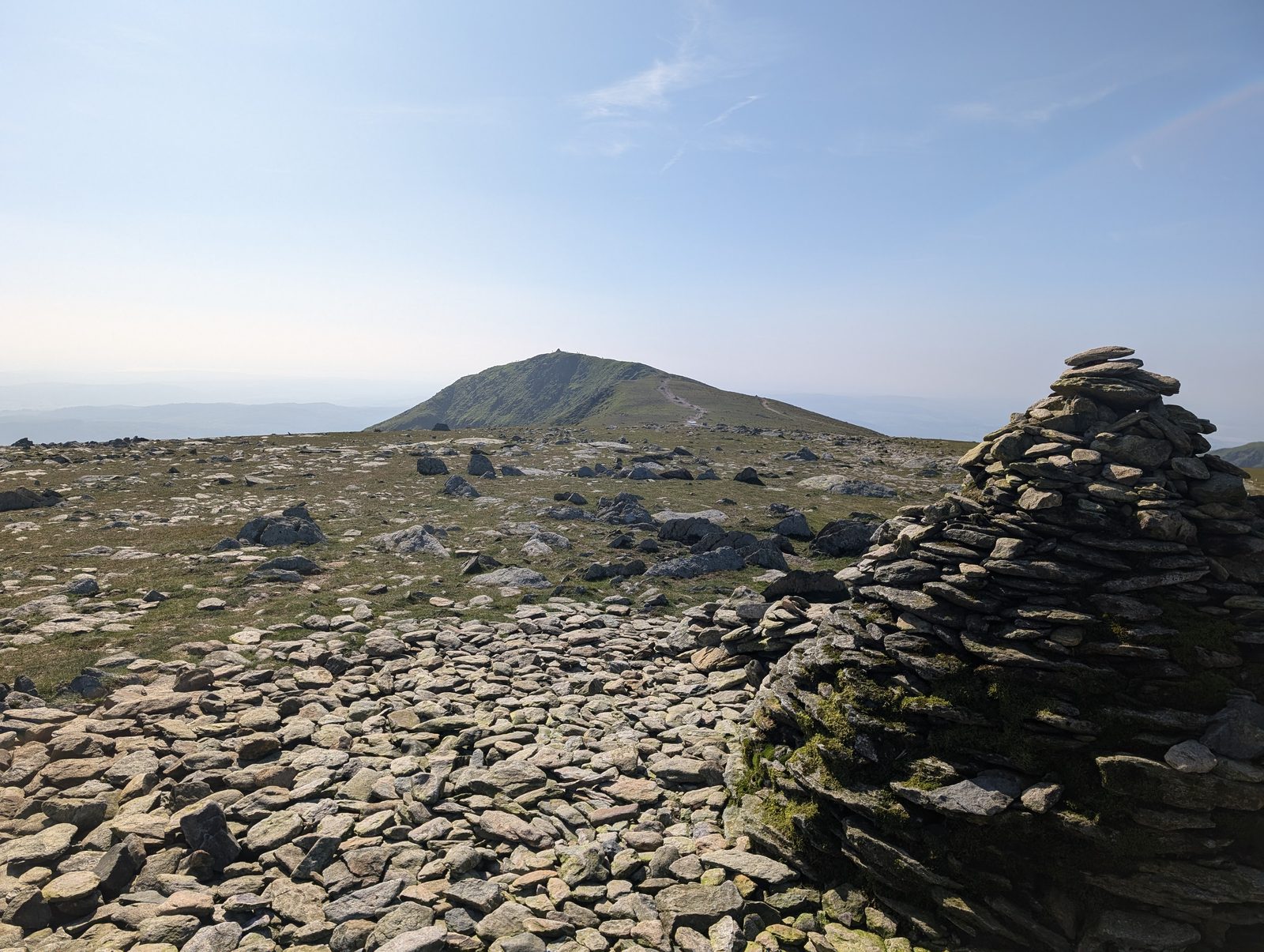 A large stone cairn on a stony, shattered summit plateau, with the next rounded top rising ahead across a shallow col