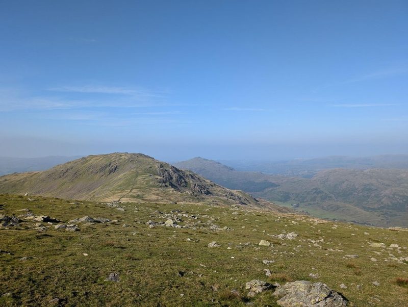 A broad grassy summit scattered with stones, with a rocky knoll ahead and a wide valley falling away to the right