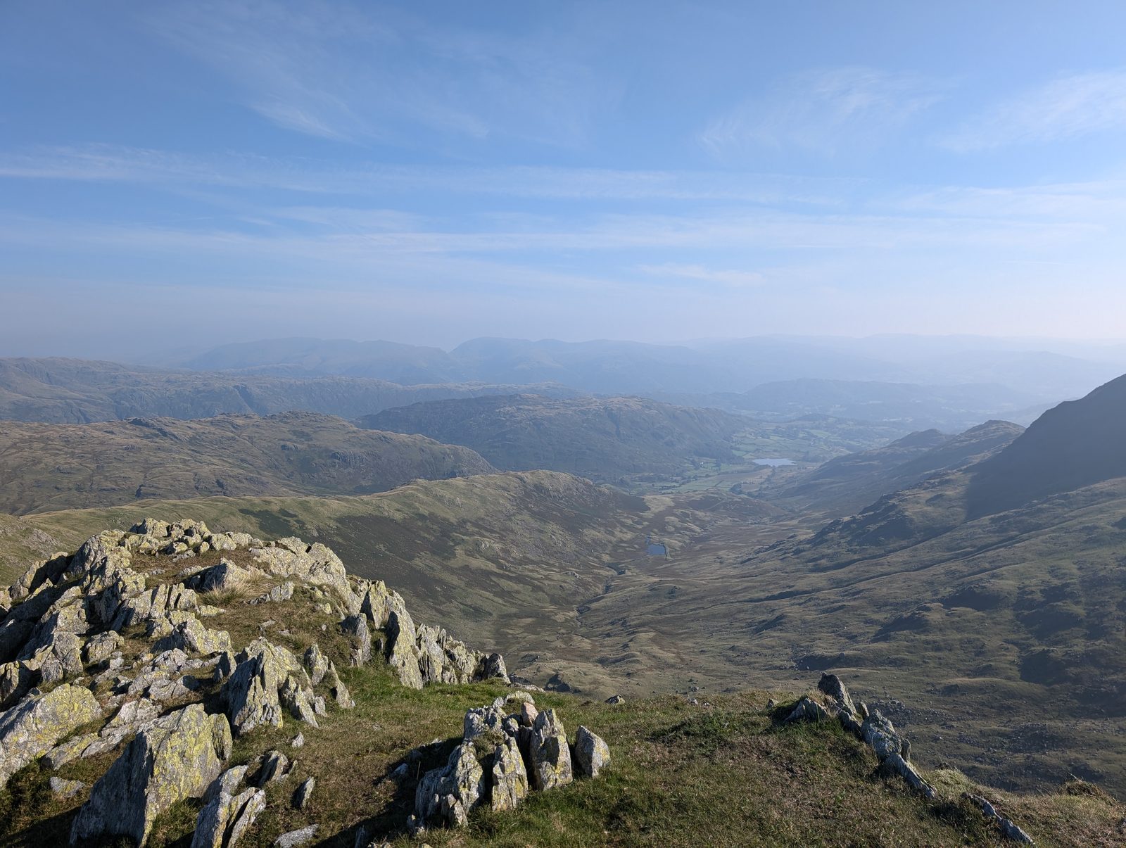 A rocky ridge crest looking down a long green valley to a small tarn and a distant lake, with hazy fells filling the horizon