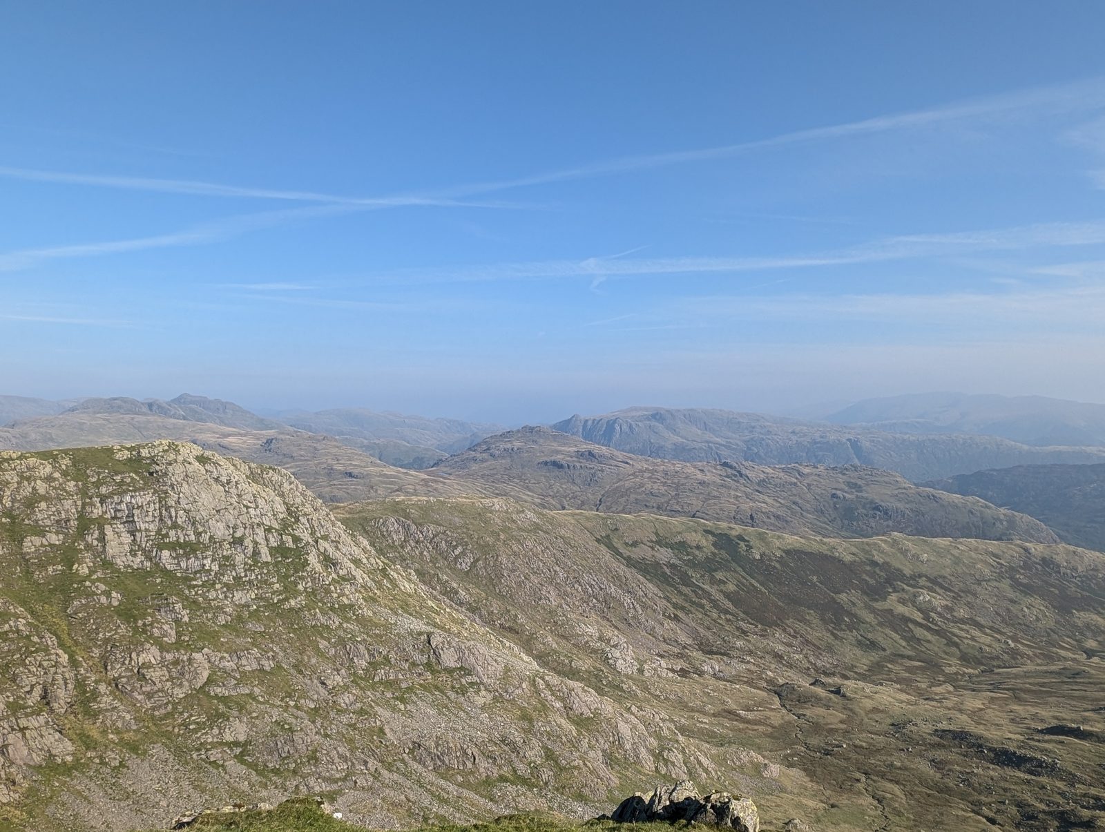 Steep crags dropping from a high shoulder into rough fellside, with rank after rank of pale hazy tops running to the horizon