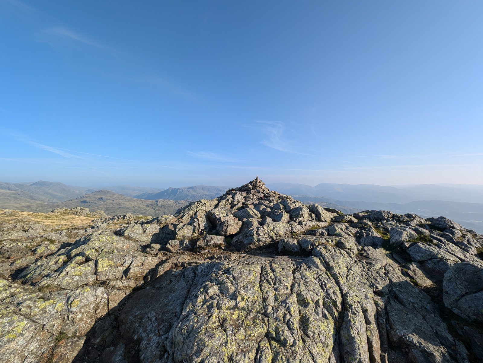 A small cairn on a summit of bare, water-worn rock slabs, with distant fells fading into haze beyond