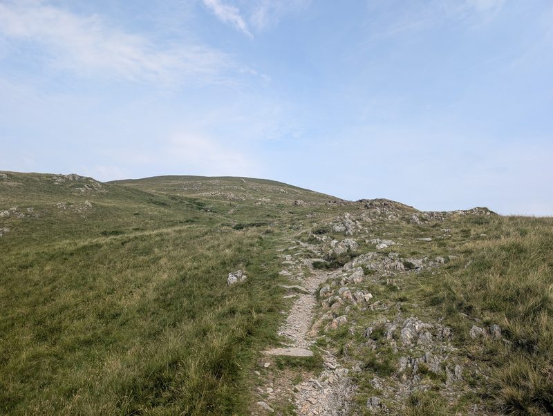 A worn stony path climbing through moor grass towards a broad, rounded grassy top under a pale blue sky