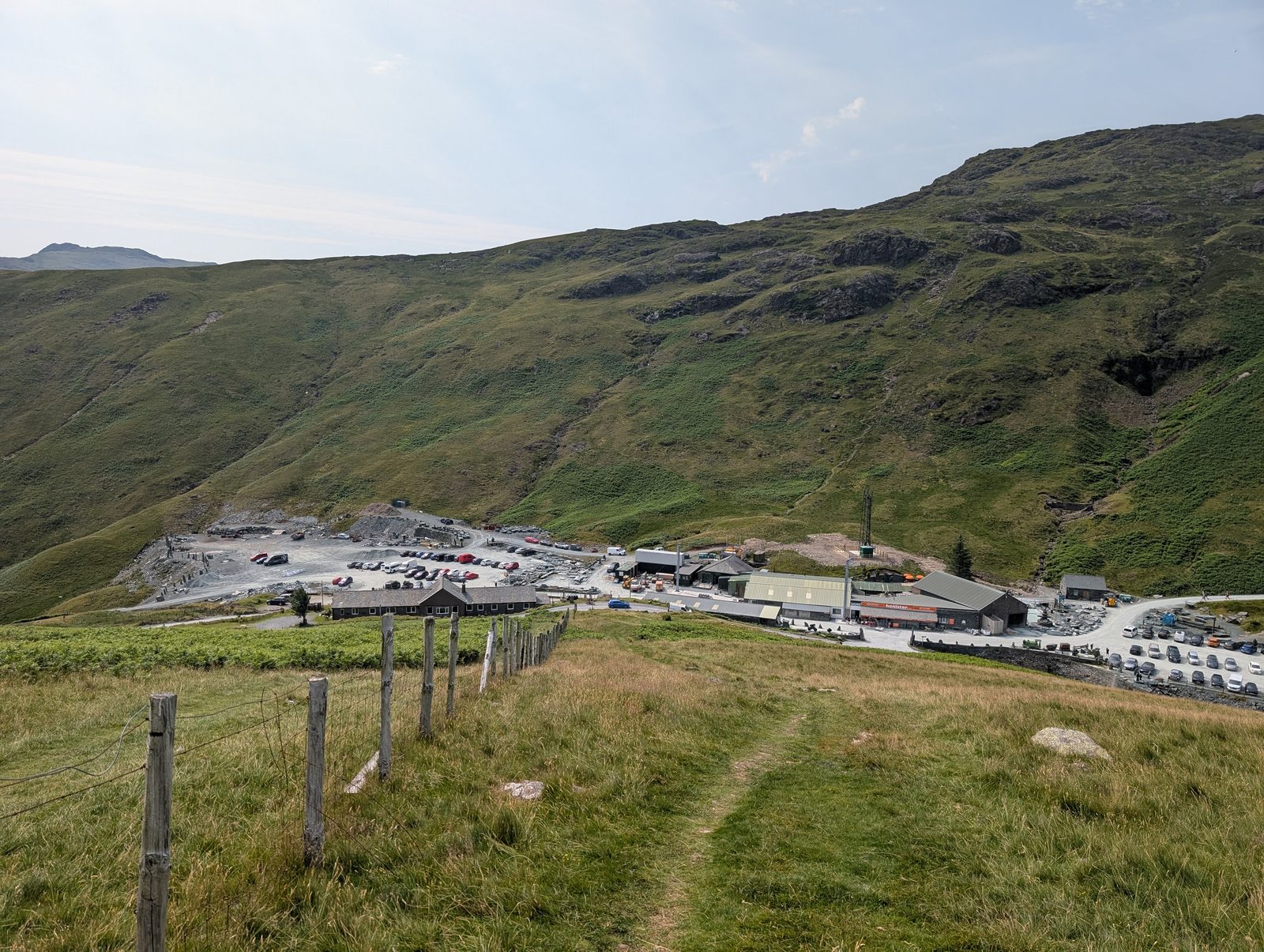 Looking down over the slate mine buildings and car park at Honister Pass, with a wire fence beside the grassy path and steep crag-strewn fellside rising behind