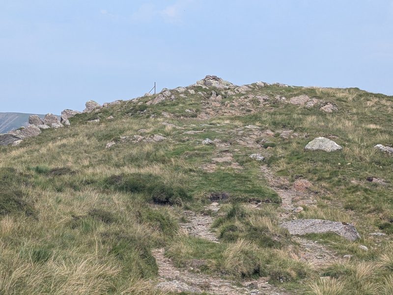 A rocky, grassy rise with a small cairn and a single fence post on the skyline, the path picking its way up through boulders and tussocks