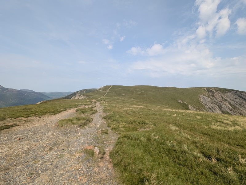 A broad grassy ridge with the path running ahead towards a rounded top, steep grey crags falling away on the right