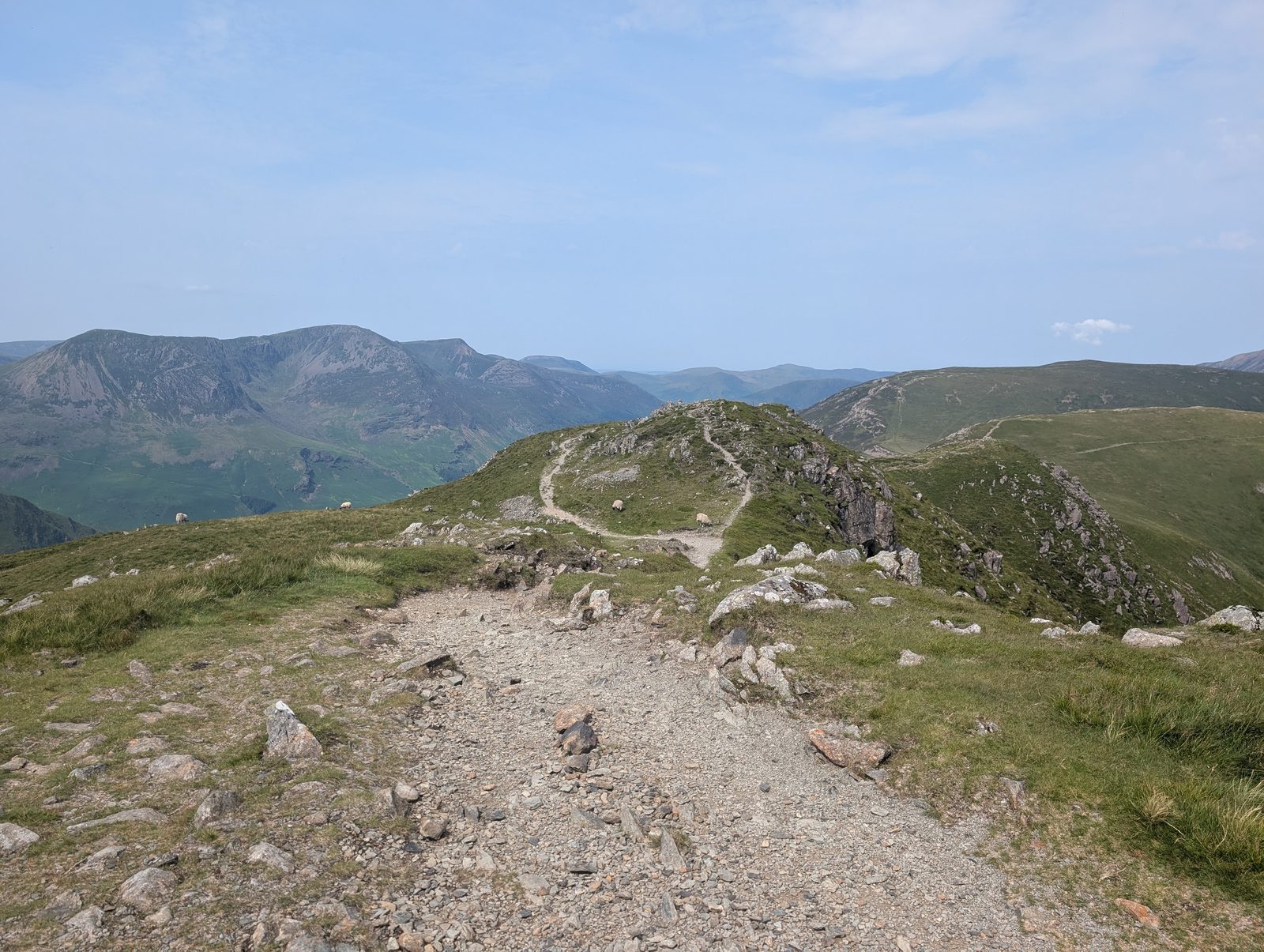 A wide stony path running along a grassy ridge with sheep grazing beside it, and a long line of steep-sided fells across the valley to the left