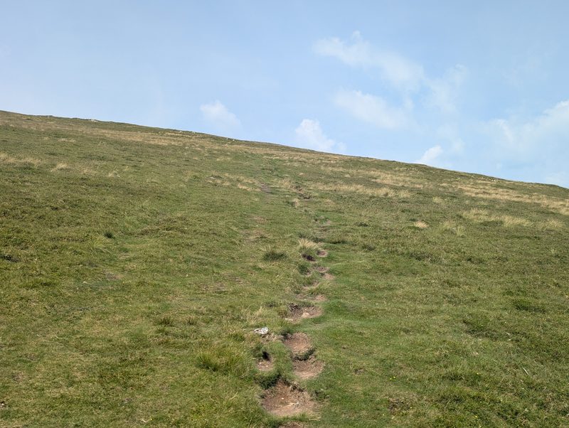 A faint, eroded path climbing straight up a wide slope of short green grass towards an empty skyline