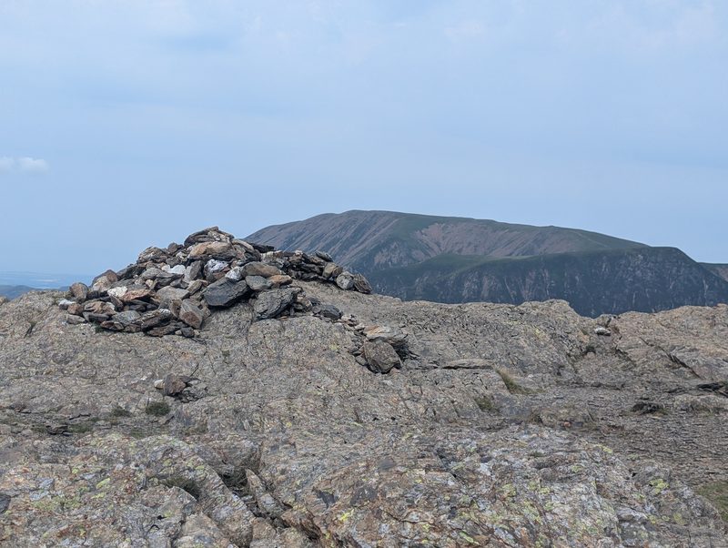 The stone cairn on Robinson's summit sitting on bare rock slabs, with a long dark ridge of fells rising across the valley behind
