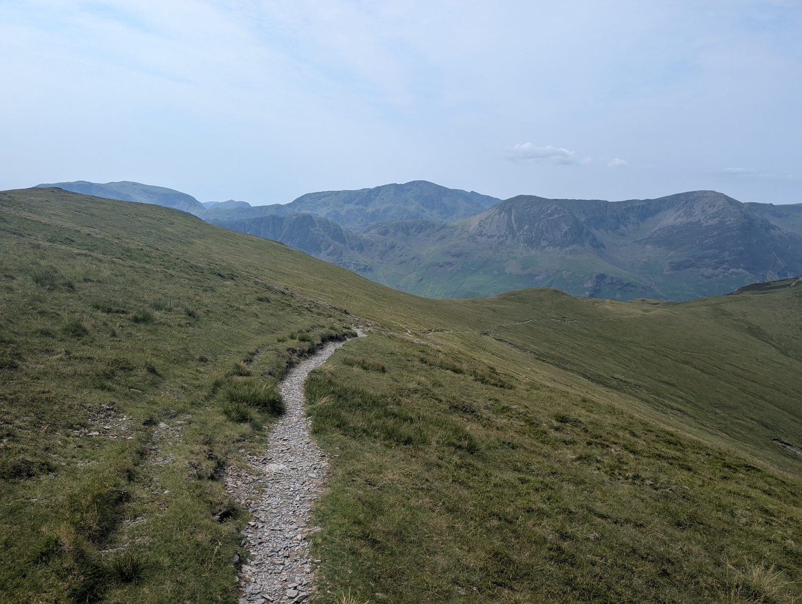 A neatly pitched stone path curving along a grassy ridge, with a wall of high craggy fells filling the skyline beyond