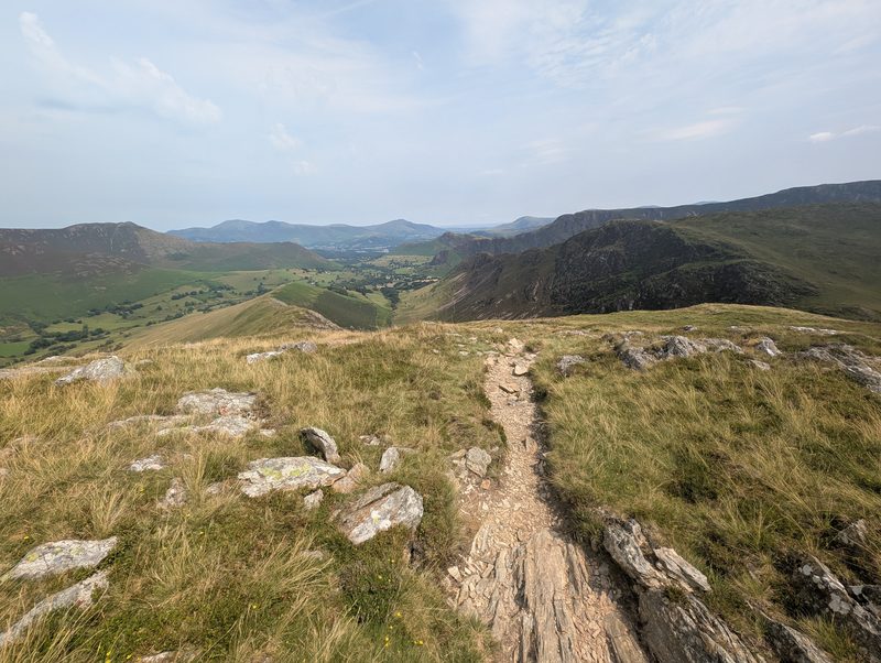 The path descending a grassy ridge between rocky outcrops, with dark crags on the right and green valley fields far below on the left