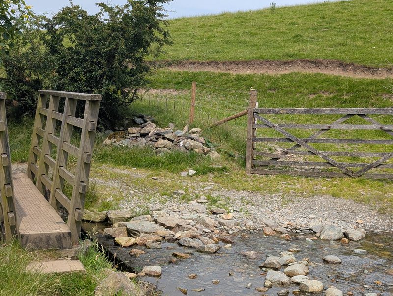 A wooden footbridge over a shallow stony beck beside a five-bar field gate, with a green pasture rising behind