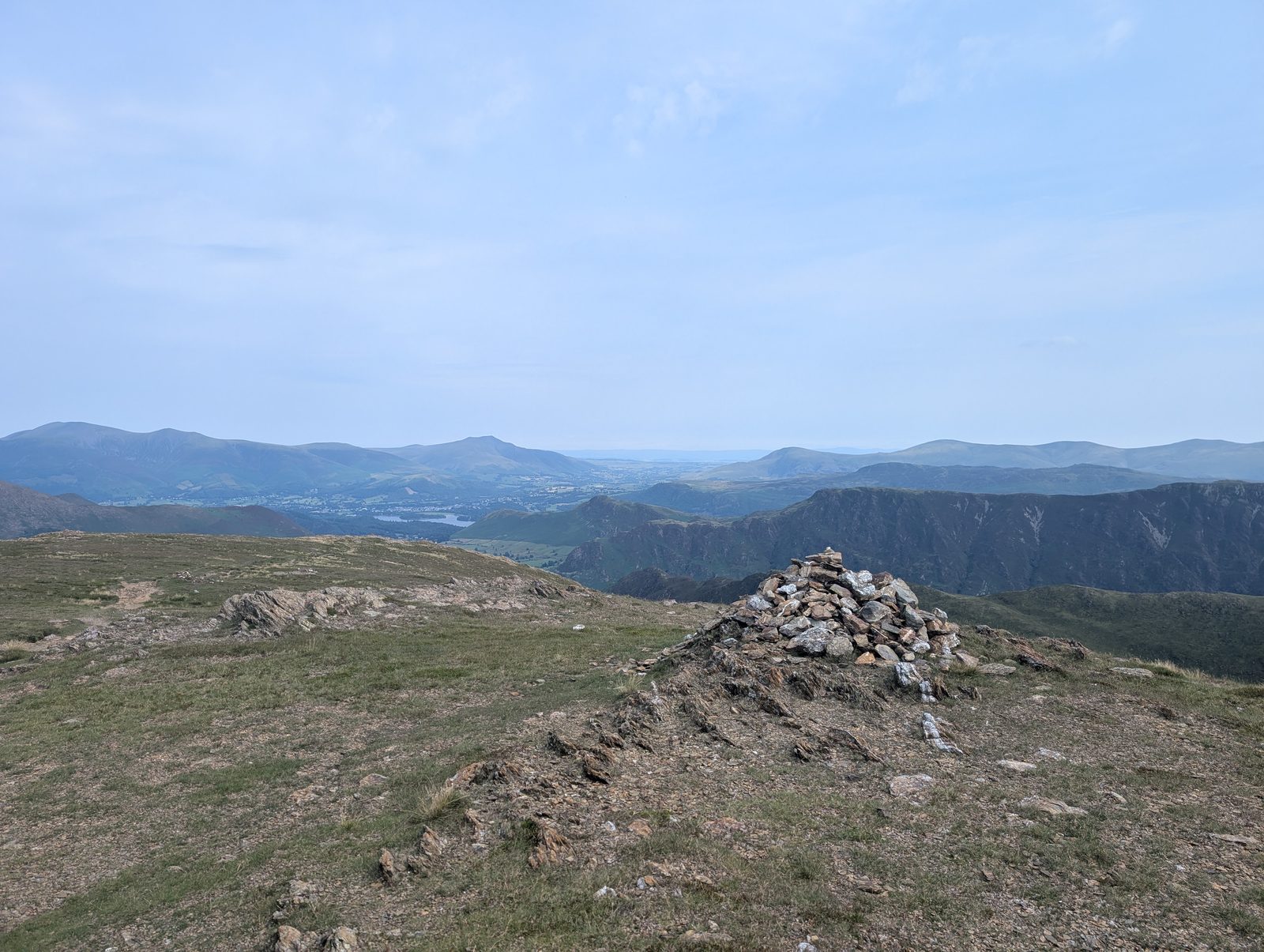 A stone cairn on a broad grassy top, looking out over Derwent Water and the rooftops of Keswick to the Skiddaw fells beyond