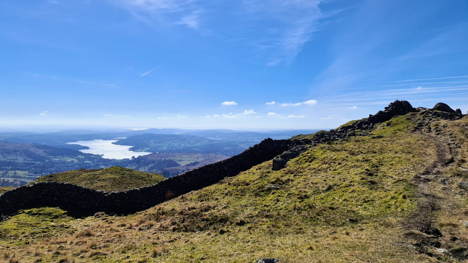 A drystone wall running along the grassy ridge crest towards rocky outcrops, with Windermere shining in the valley far below