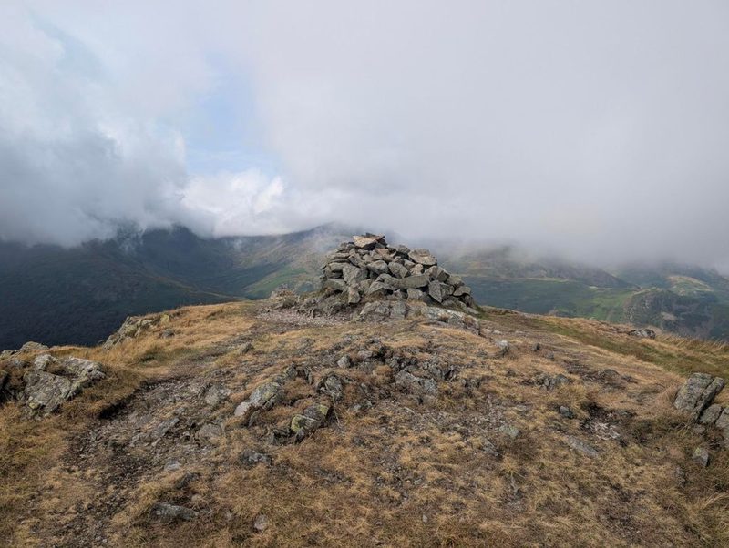 A large cairn of angular blocks on a grassy top, with cloud pouring over the high fells across the valley behind