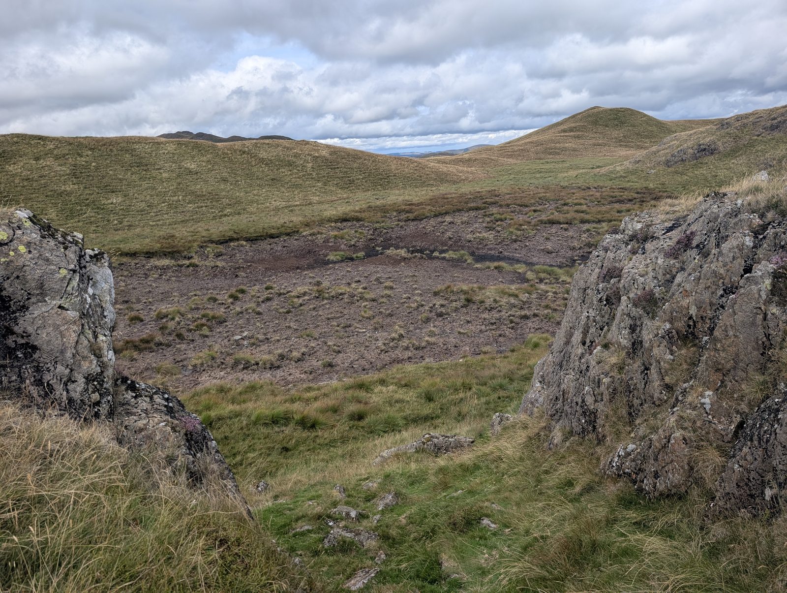 A rocky notch on a heathery ridge looking across a wide peaty hollow to smooth grass tops beyond, under a sky of broken cloud