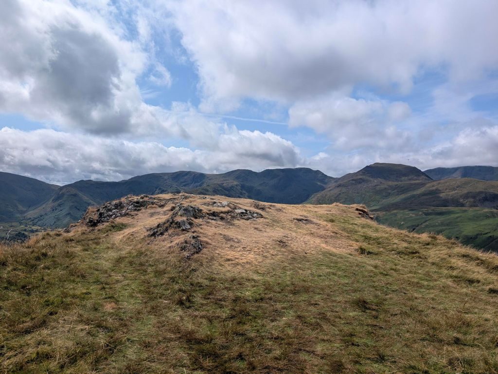 A broad, rough grassy summit with a low rocky outcrop, looking across a deep valley to a long line of high fells under bright cloud