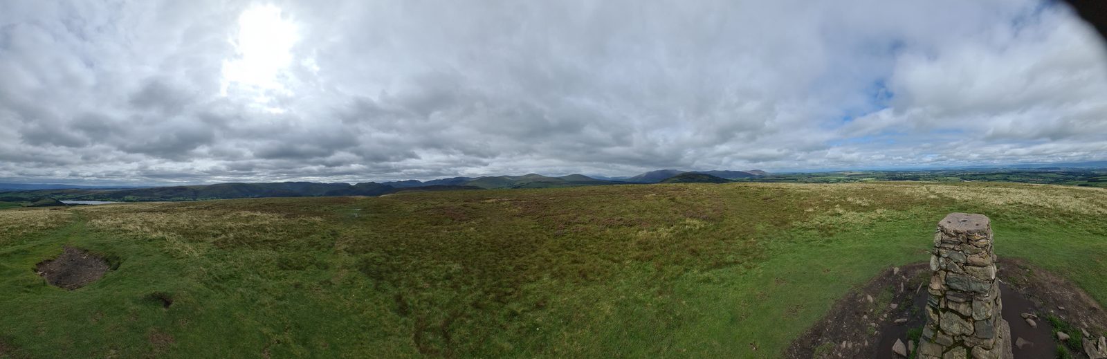 A wide view from the broad grassy dome of Little Mell Fell, its stone trig column at the right, heather moor in the foreground and distant fells under heavy cloud