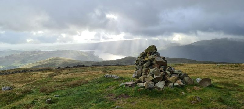 A shallow rocky beck running through a green valley between drystone walls, with scattered trees and low grassy fells under a broken sky