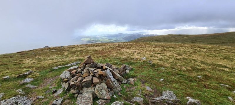 A tall stone cairn on a grassy summit, with shafts of light breaking through heavy cloud over Ullswater and the fells beyond